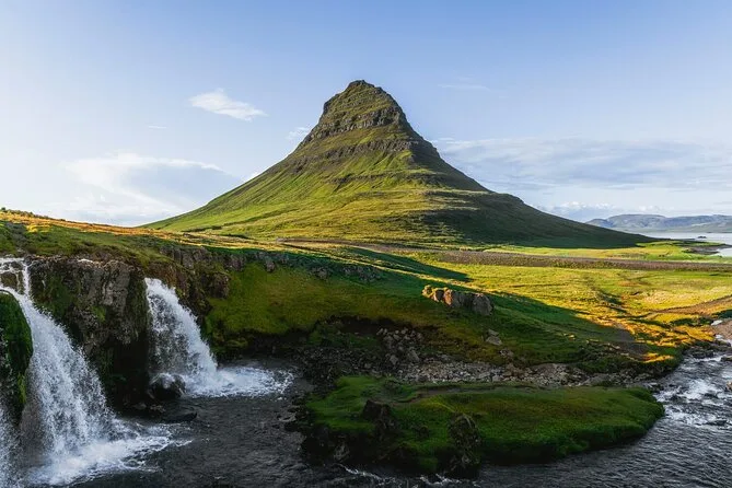 Kirkjufell under a clear sky, seen above the twin drops of Kirkjufellsfoss and a mossy riverbank