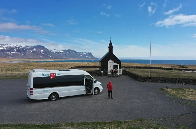 Ein weißer Minibus an der schwarzen Kirche von Búðir, daneben steht ein Gast in Rot