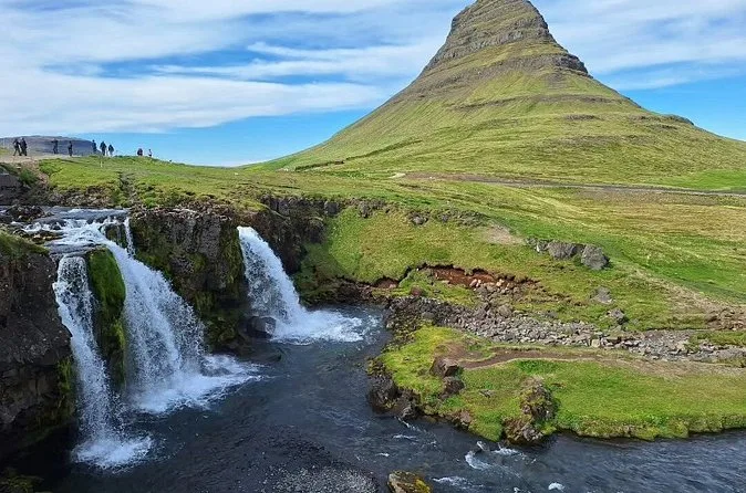 Kirkjufellsfoss in two streams below the green slope of Kirkjufell, with a few visitors on the ridge to the left